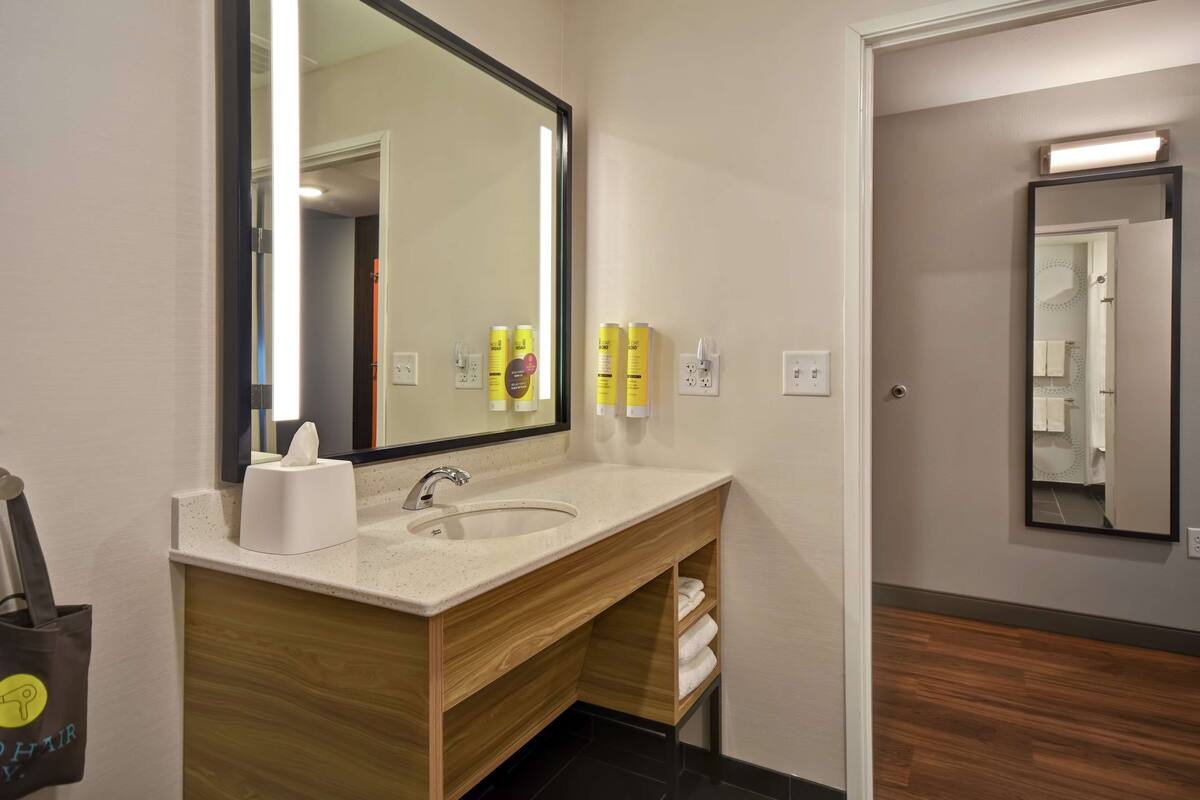 Un baño moderno con un espejo grande, un lavabo y armarios de madera. Hay artículos de aseo en la encimera y un perchero debajo del lavabo.