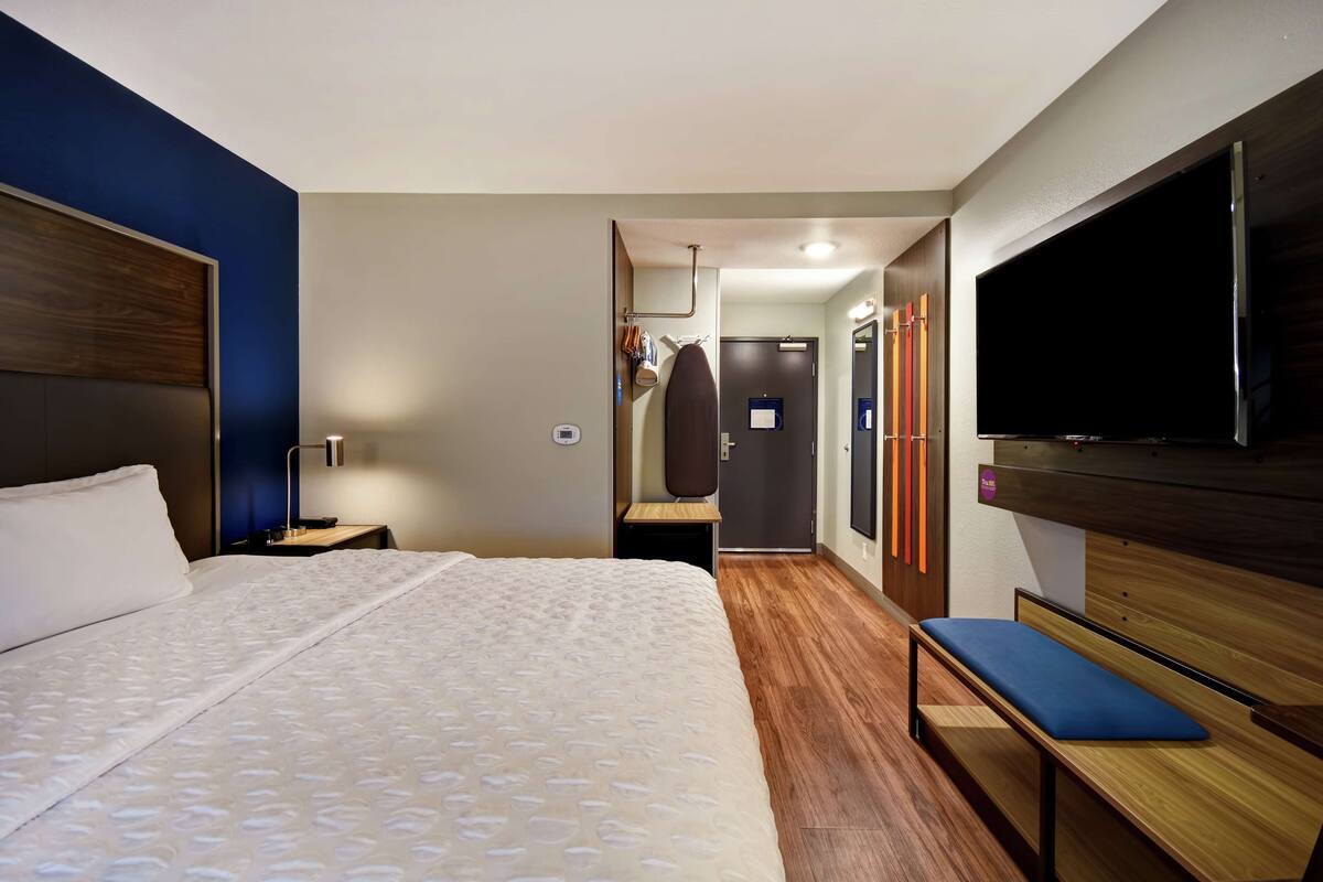 Una habitación de hotel con una cama grande, televisión, banco y espejo.