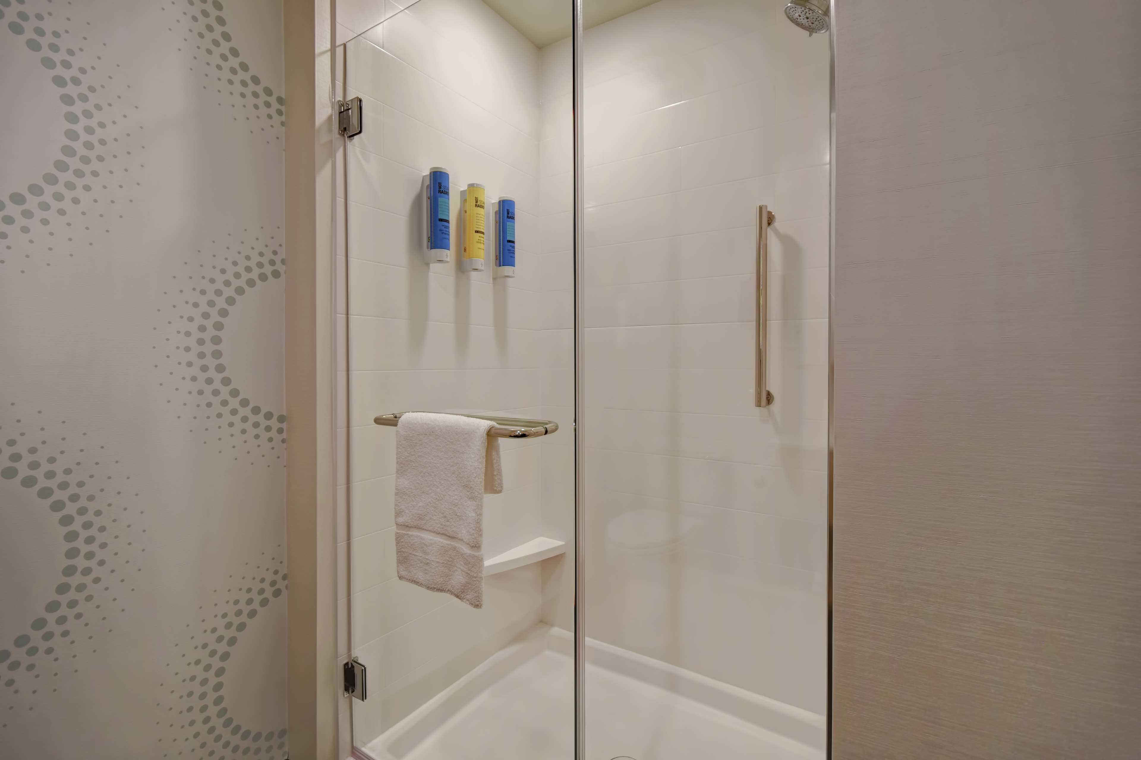Zona de ducha con cerramiento de vidrio, pared con azulejos blancos, un perchero con toalla blanca y tres artículos de tocador en la pared.