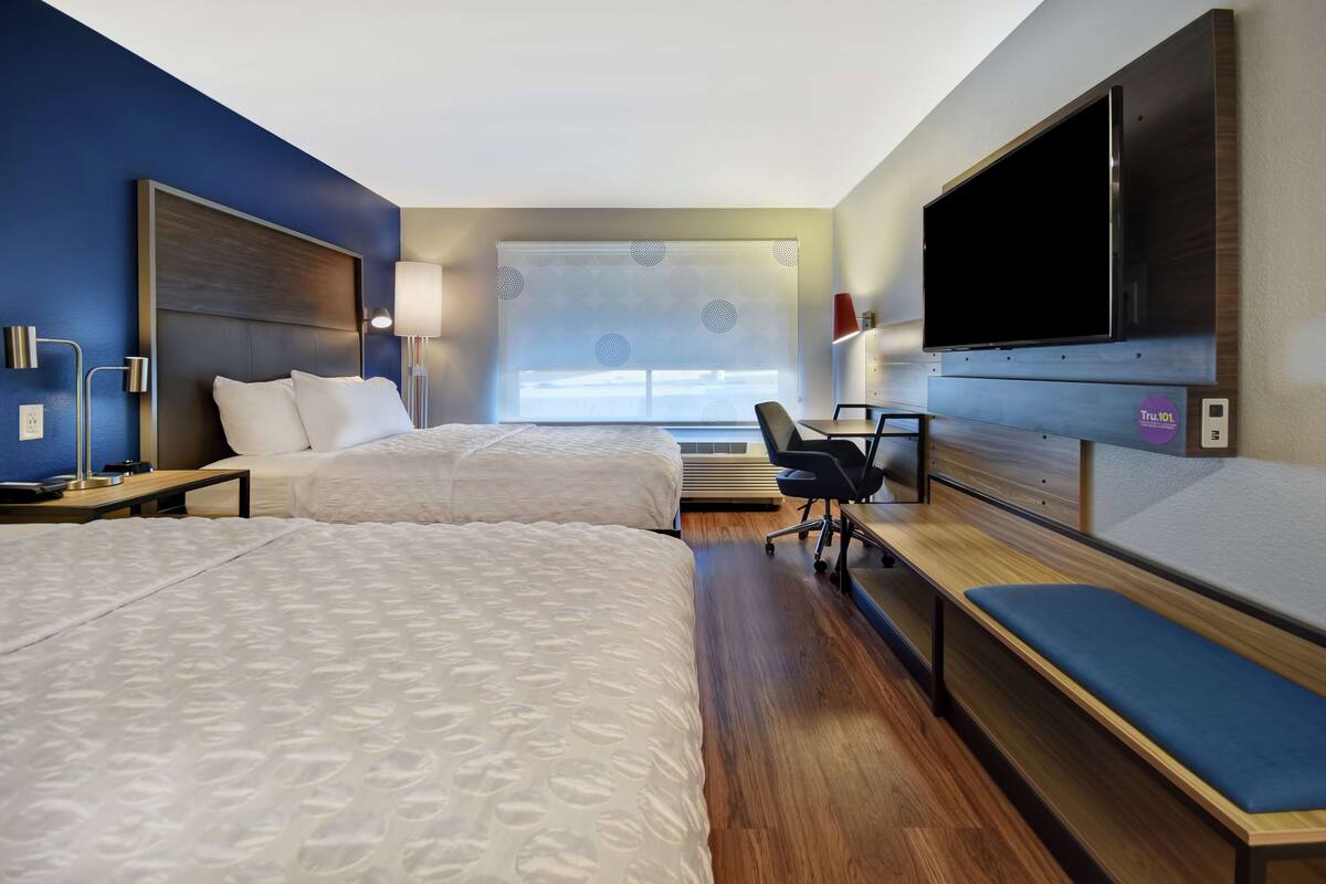 Habitación de hotel con dos camas, un escritorio, una silla, televisión y un ventanal con persianas.