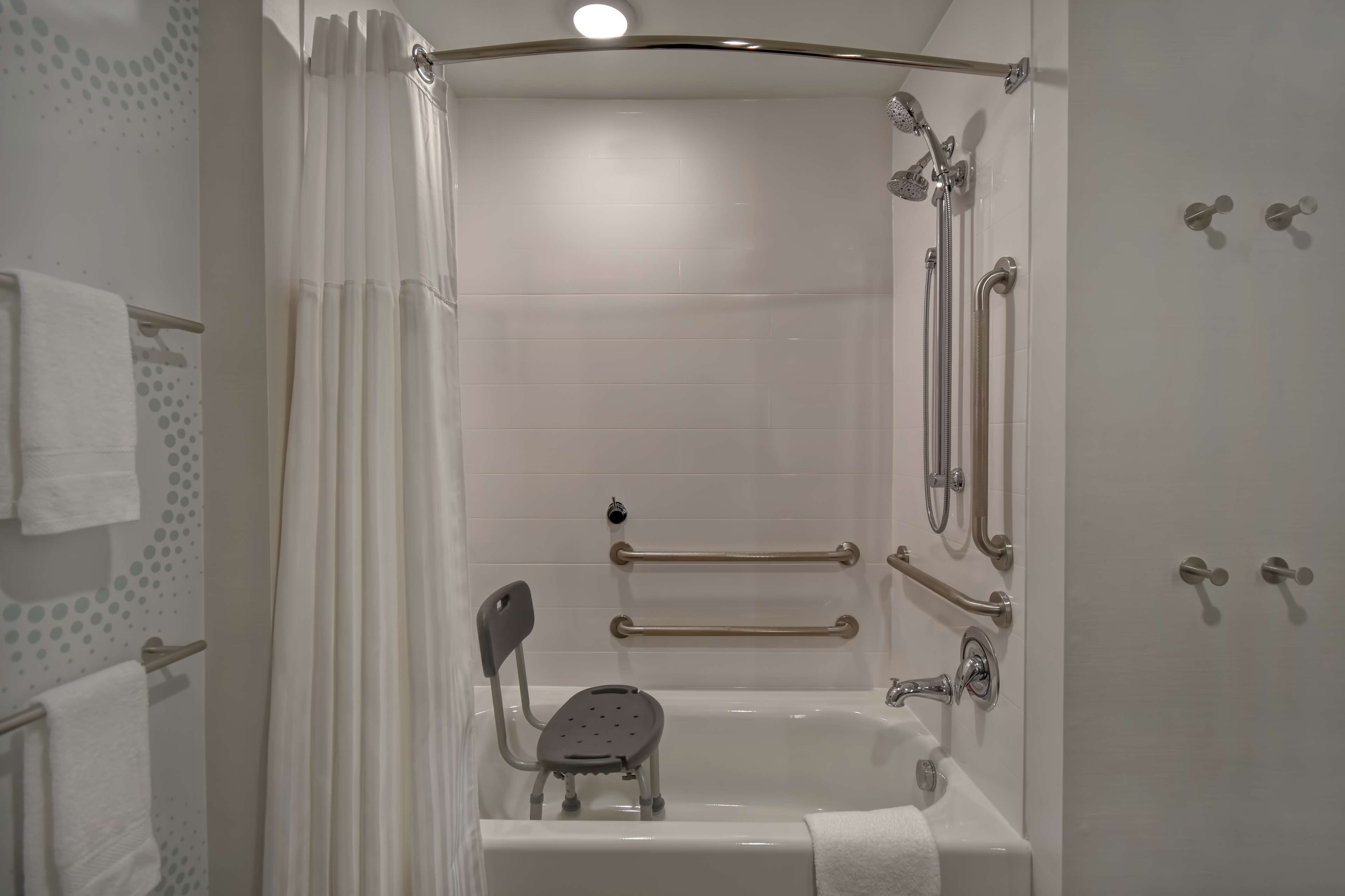 Un baño moderno con cabina de ducha a ras de suelo, barras de apoyo, cabezal de ducha de mano y un taburete pequeño.