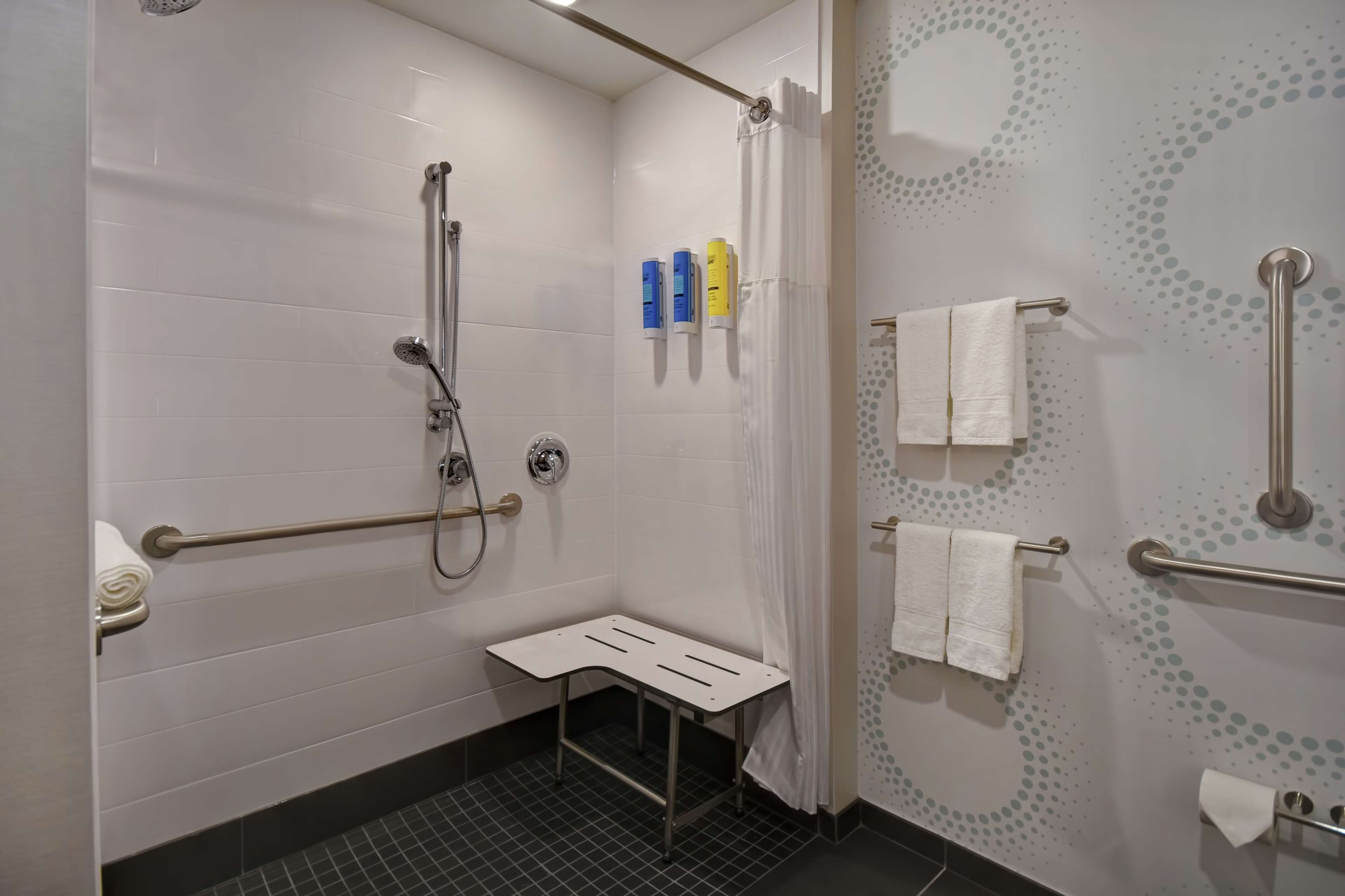 Ein modernes Badezimmer mit weiß gefliesten Wänden, einer Dusche inklusive Duschbrause, einer Haltestange und einer kleinen Sitzbank. Zwei Handtuchhalter mit weißen Handtüchern sowie ein Toilettenpapierhalter sind vorhanden.