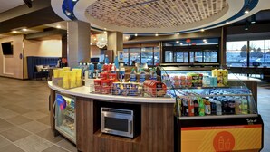Una estación autoservicio de bebidas con diversas opciones de bebidas y snacks, una máquina de café y vistas al estacionamiento a través de amplios ventanales.