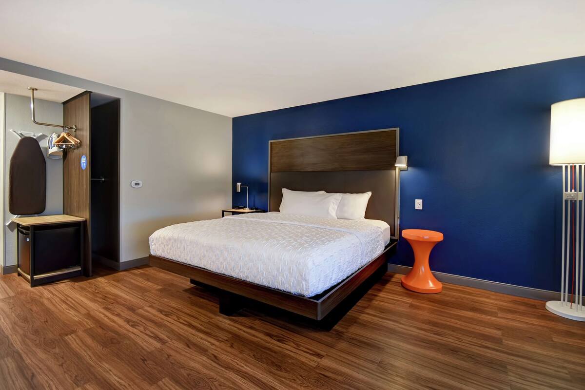 Una habitación de hotel moderna con una cama grande, piso de madera, pared azul como detalle, un taburete naranja pequeño y un espejo con perchero.