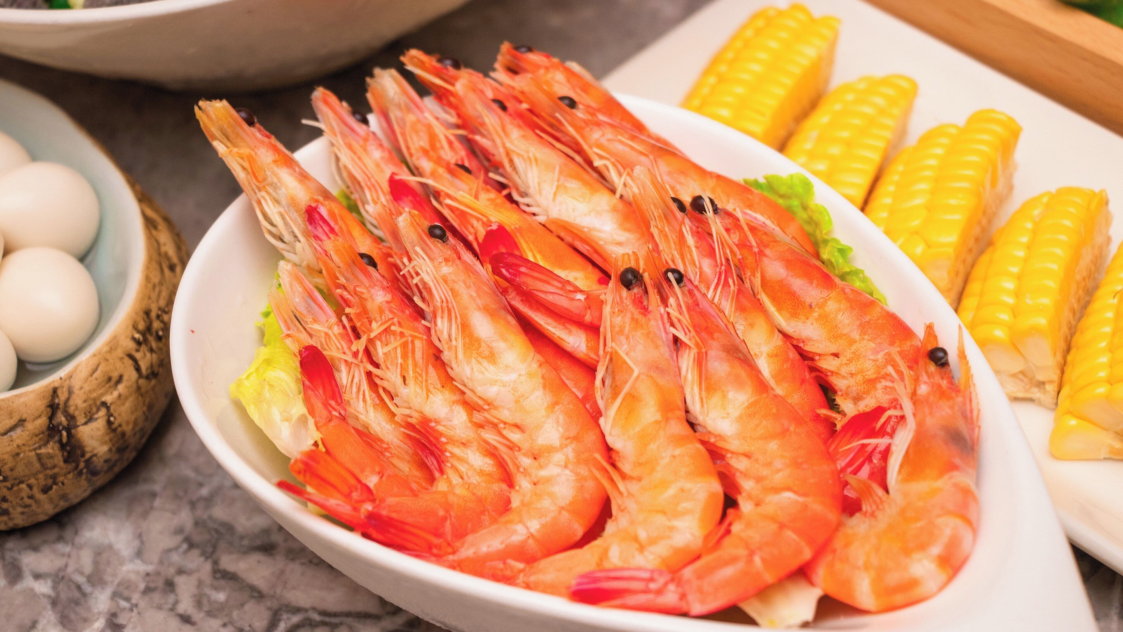 A plate of cooked shrimp with corn on the cob and a bowl of eggs.