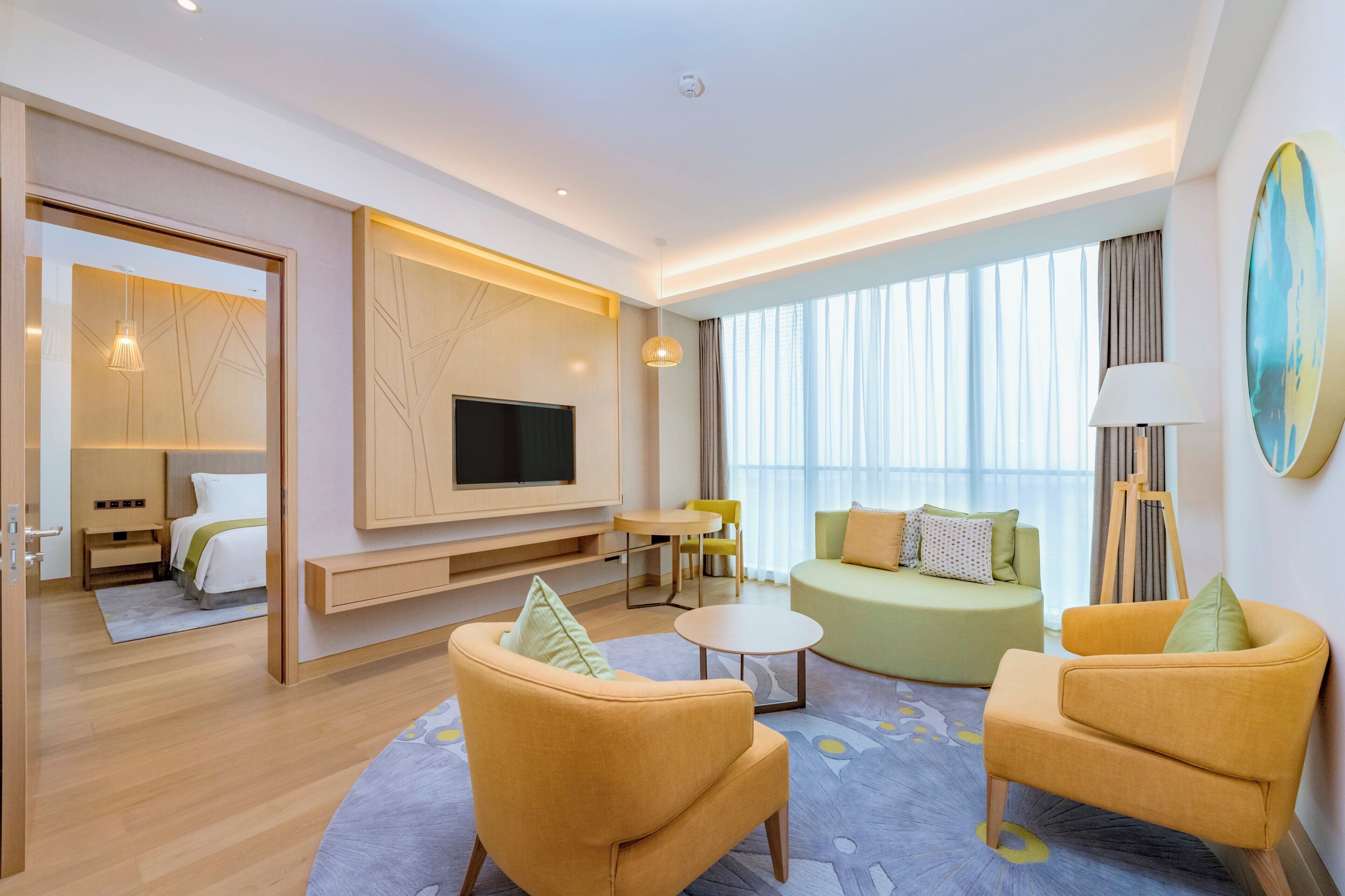Una habitación de hotel moderna con una televisión de pantalla plana, dos sillones amarillos, un sofá verde, una mesita redonda y un ventanal con cortinas.