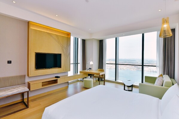 Ein modernes Hotelzimmer mit einem großen Bett, einem Schreibtisch, einem Sessel, einem an der Wand befestigten Fernseher und Blick auf die Stadt durch große Fenster.