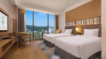 Habitación de hotel con dos camas, un escritorio y un amplio ventanal con vista a la ciudad.