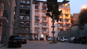 A multi-story residential building with balconies, parked cars, and a tree in the foreground.