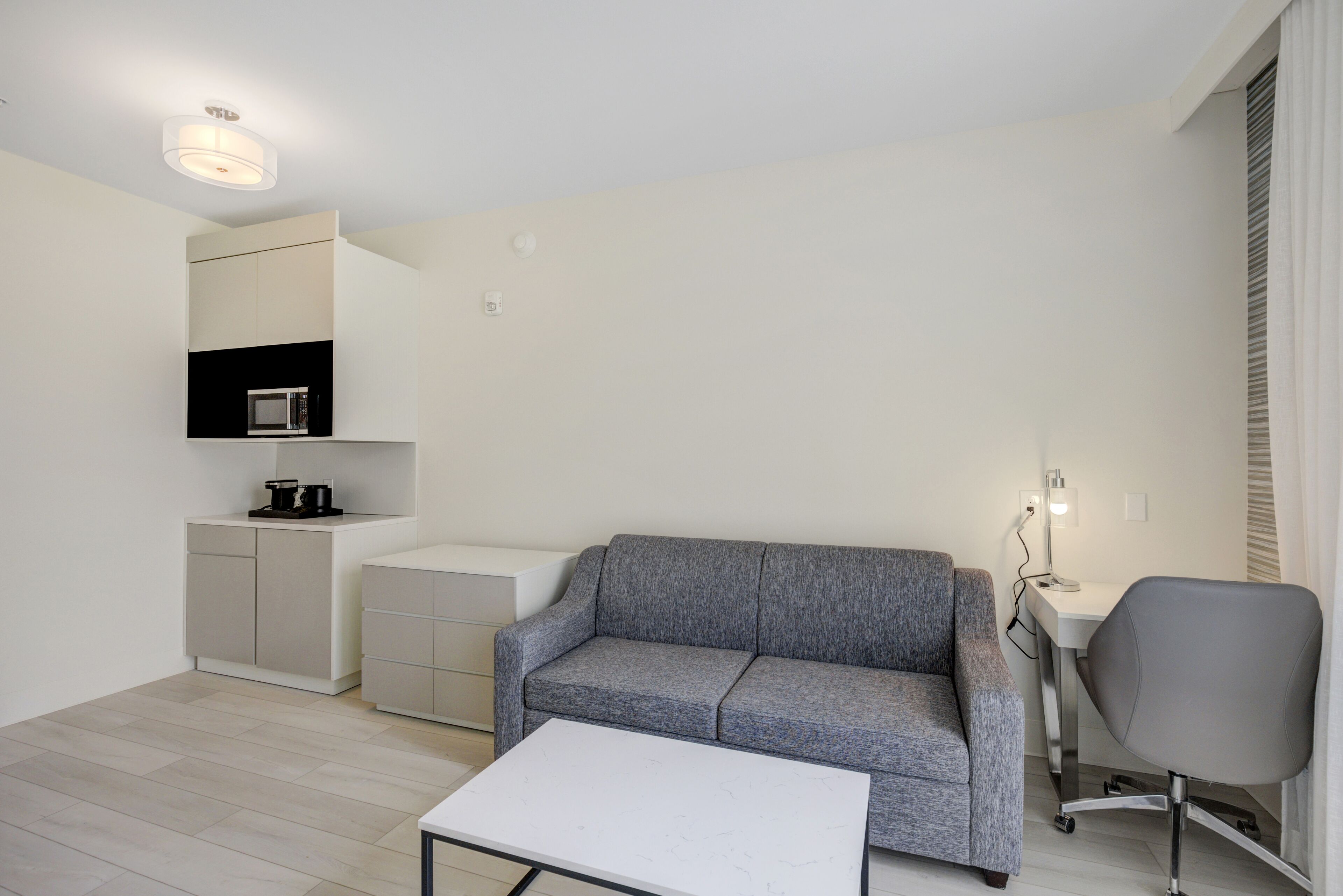 Un salon moderne avec un canapé gris, une table basse blanche, un fauteuil de bureau gris, un petit coin cuisine équipé d’un micro-ondes et un luminaire blanc au plafond.