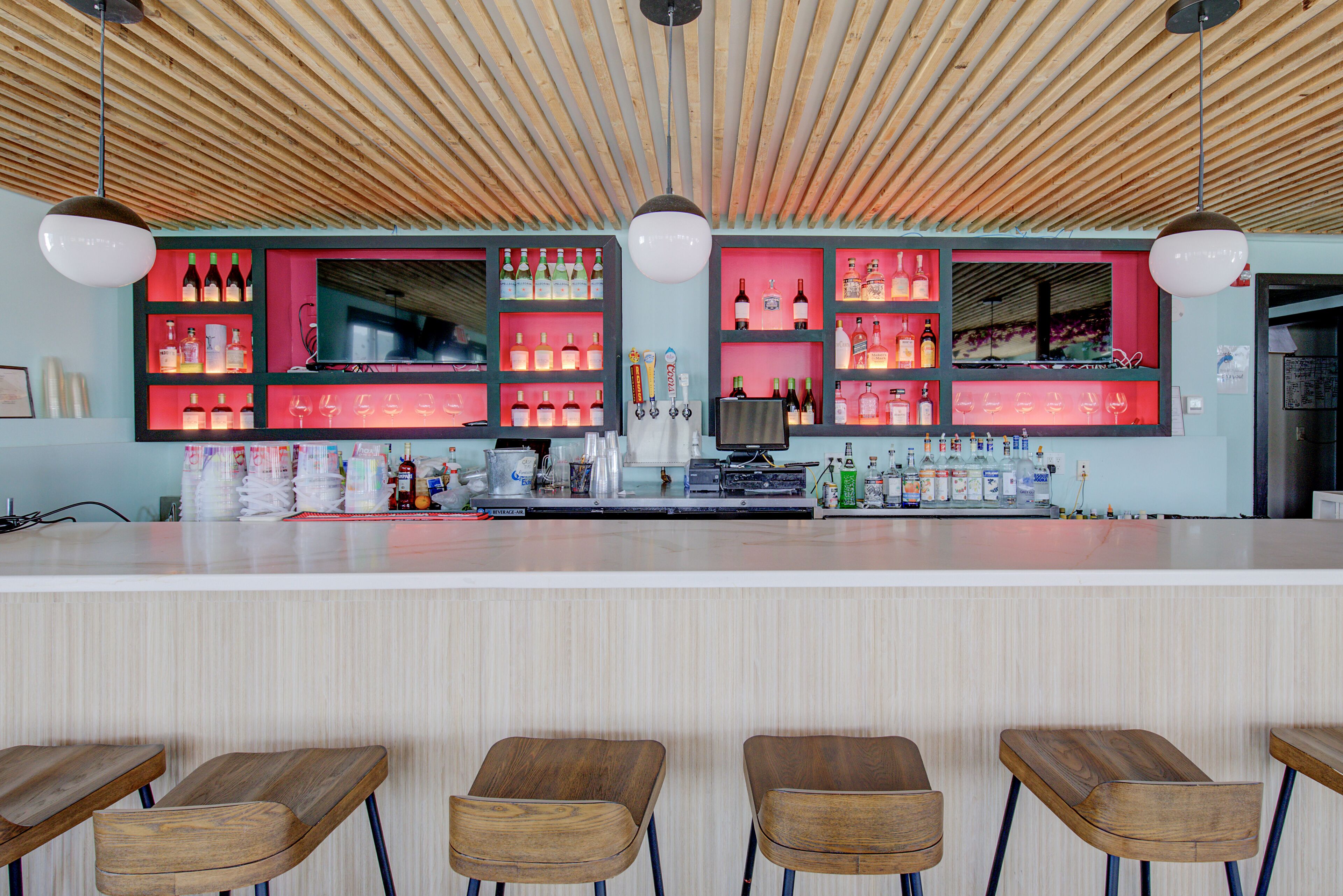 A bar area with wooden ceiling, shelves with bottles, and bar stools.