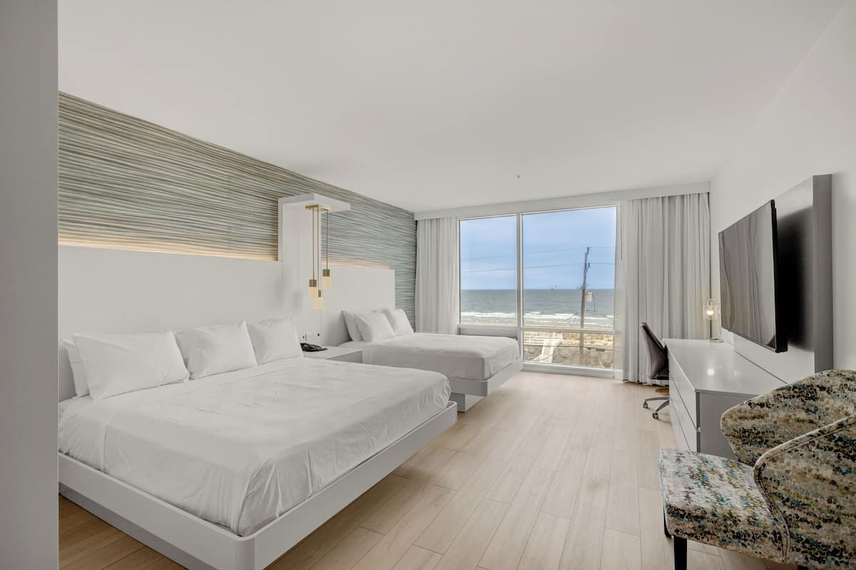 A modern hotel room with two beds, a large window with a view of the sea, a flat-screen TV, and a desk with a chair.