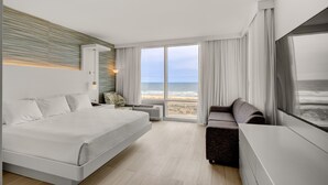 A modern hotel room with a large bed, a sofa, a TV, and a view of the beach.