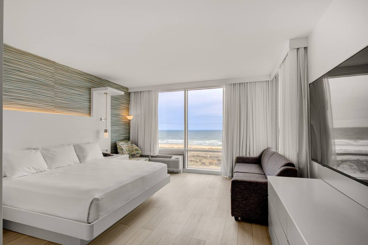 A modern hotel room with a large bed, a sofa, a TV, and a view of the beach.