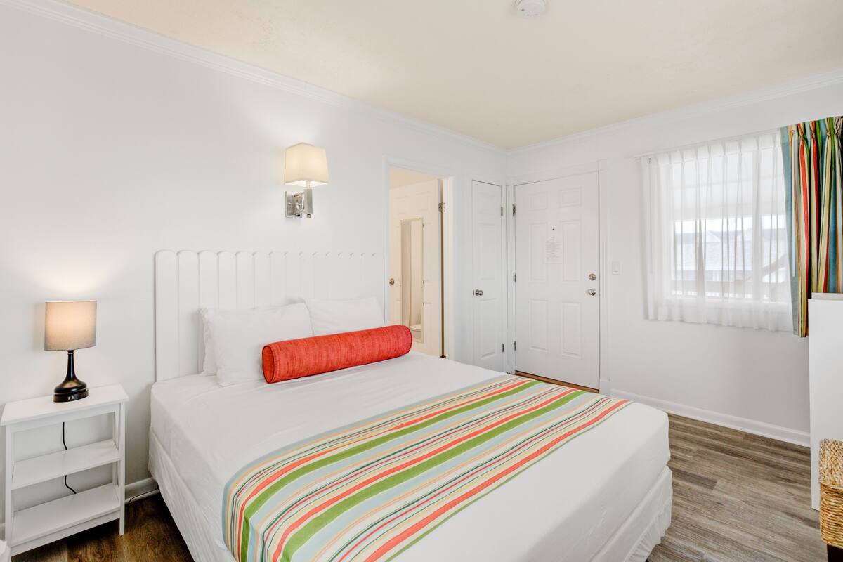 A hotel room with a bed, a nightstand, a lamp, a door, and a window with curtains.