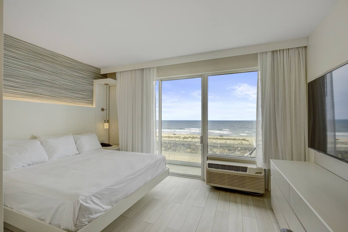 A hotel room with a bed, a large window with a view of the beach, and a flat-screen TV.