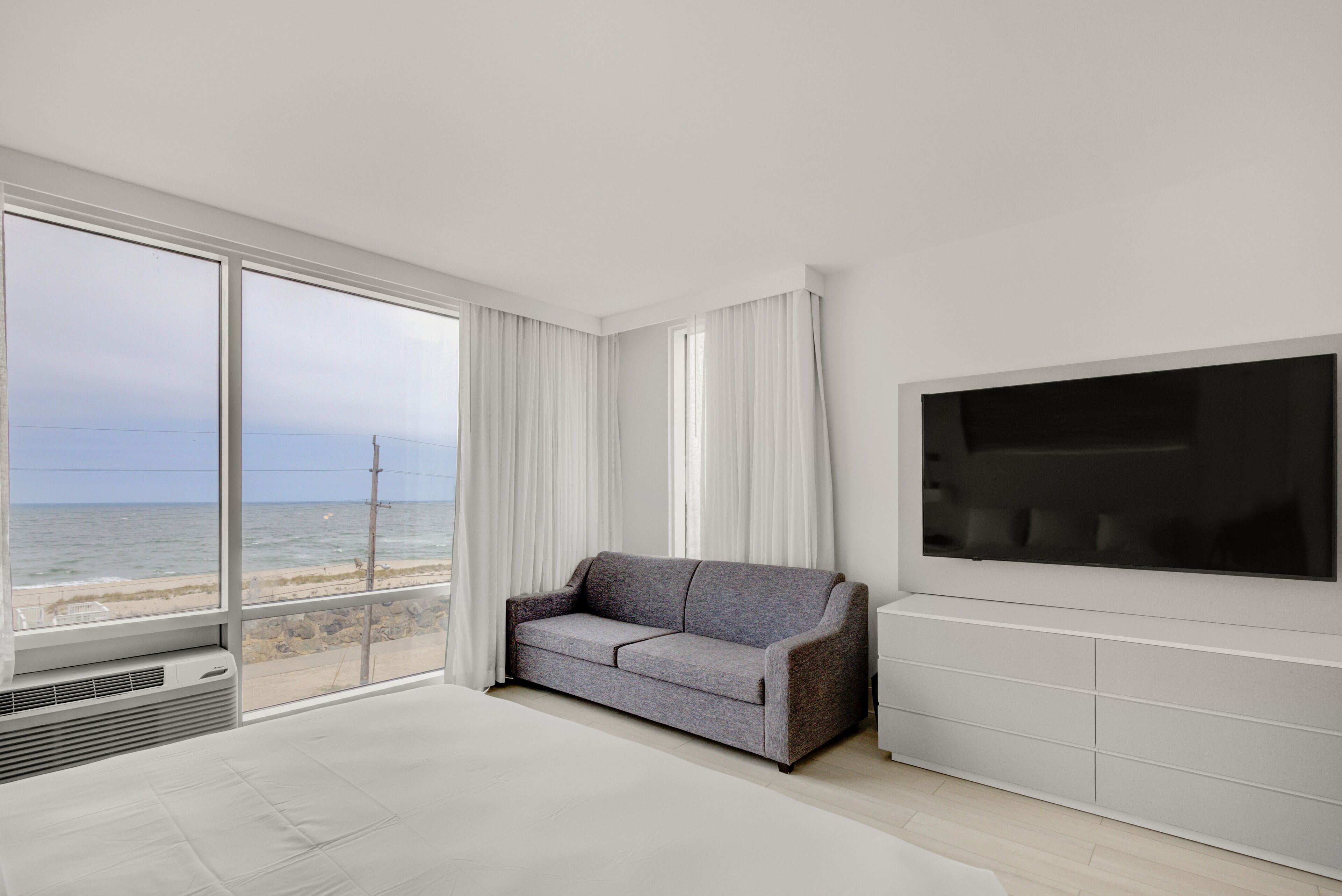 Ein modernes Hotelzimmer mit großem Fenster und Blick auf den Strand, einer grauen Couch, einem Flachbildfernseher und einem minimalistischen Design.