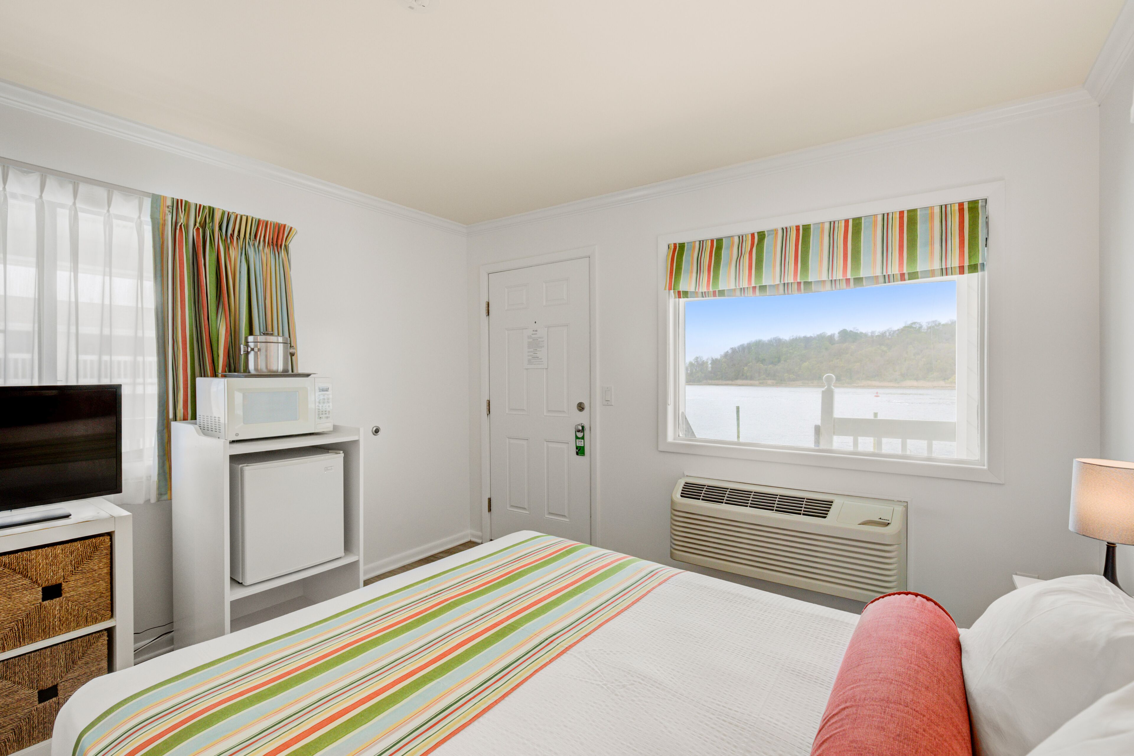 A hotel room with a bed, a TV, a microwave, a small fridge, a window with a view of a body of water, and a door with a sign.