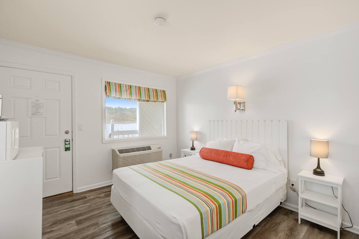 A bedroom with a bed, white walls, a window with a view, a door, and a nightstand with a lamp.