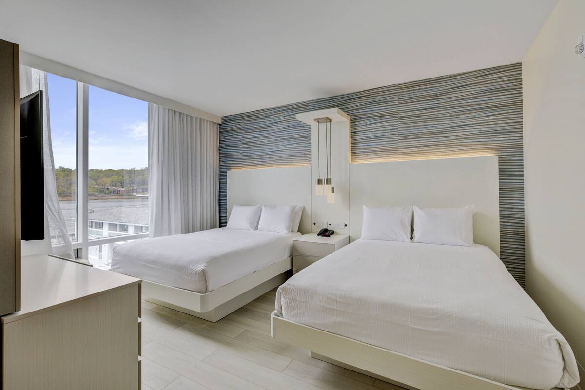 A hotel room with two beds, a large window with curtains, a flat-screen TV, and a modern light fixture.