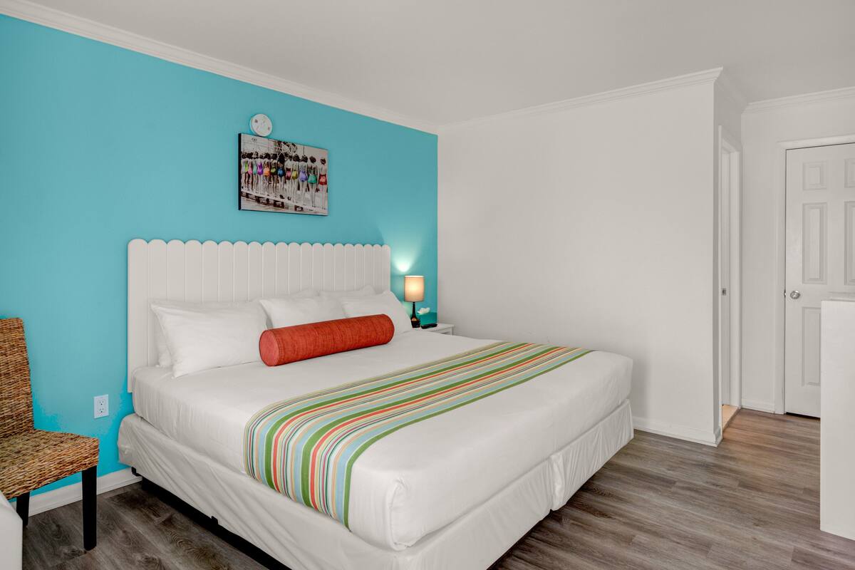 A bedroom with a large bed, a nightstand with a lamp, a wicker chair, and a door.