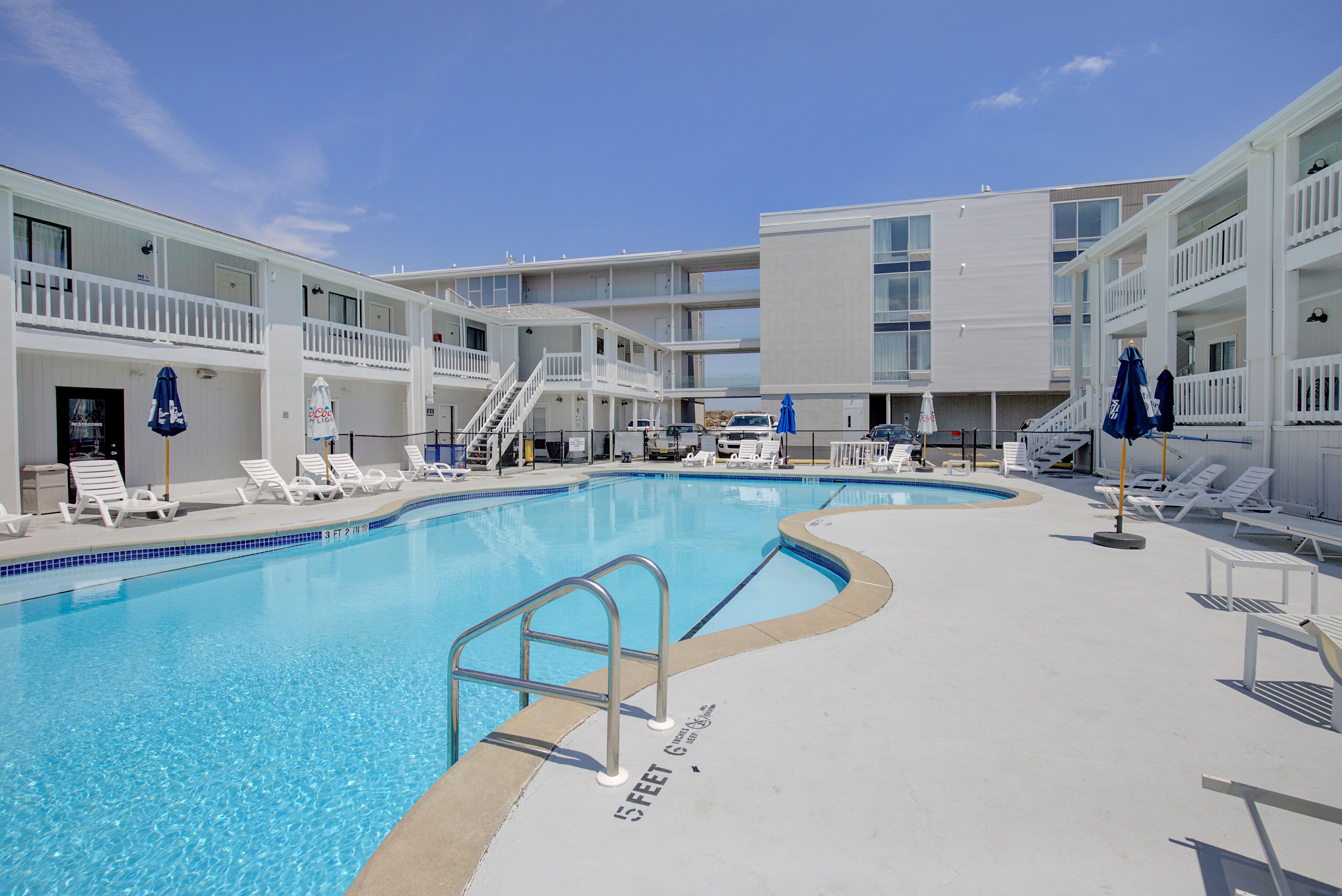 A swimming pool with white lounge chairs and umbrellas, surrounded by a multi-story building with balconies.