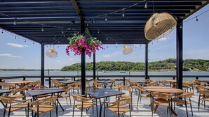 Un patio exterior con mesas y sillas, una pérgola y vistas a un lago y follaje verde.