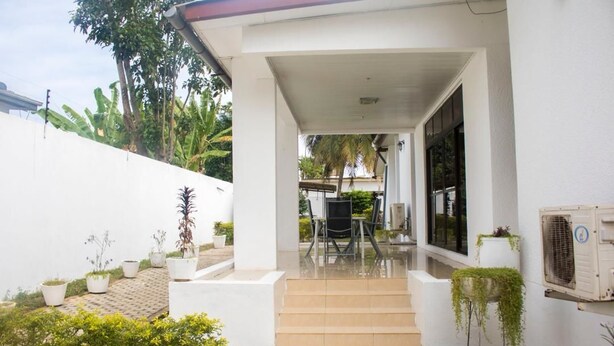 A covered outdoor area with steps leading to a glass door, surrounded by potted plants and a white wall. - Sara's place (Tema)