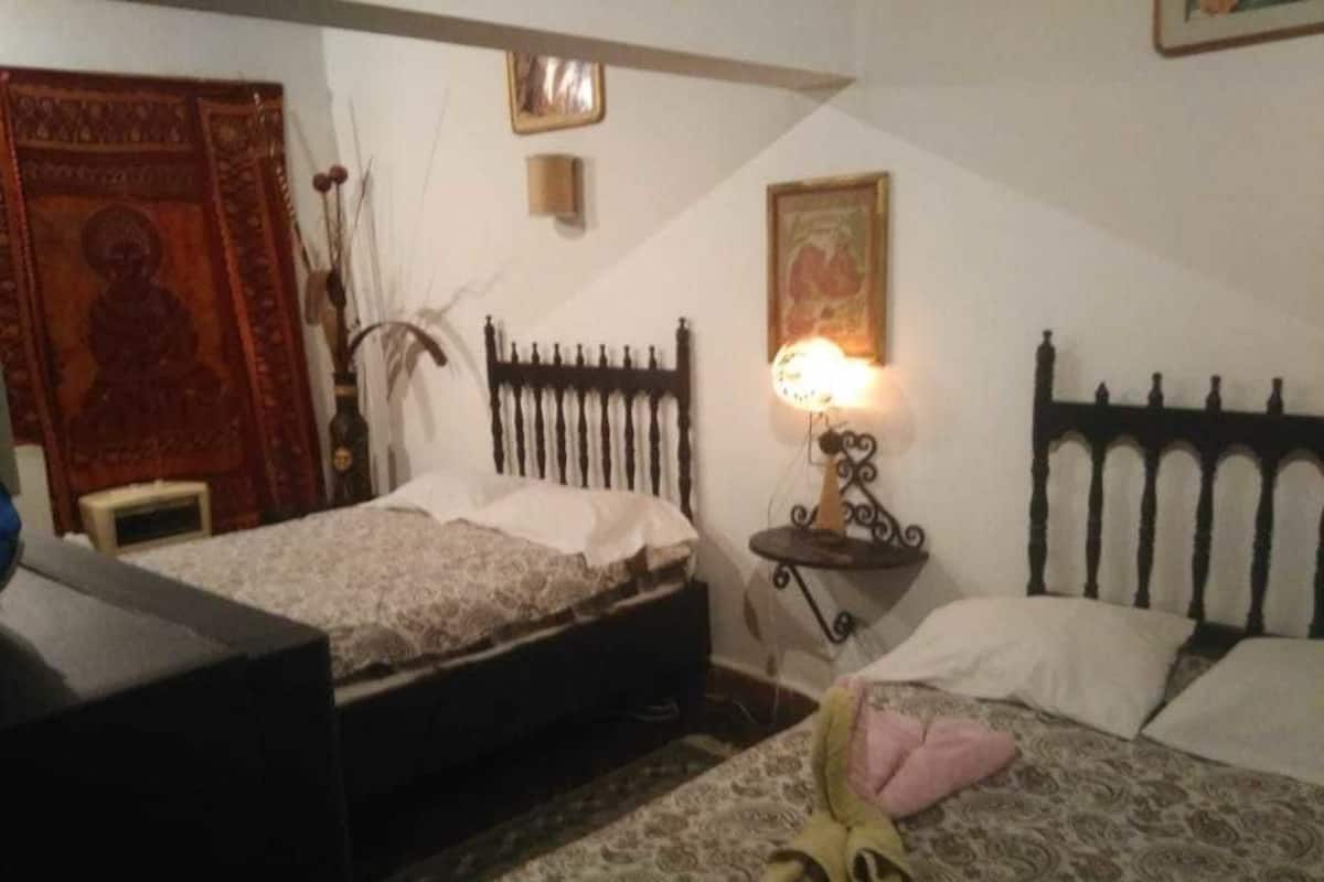 Un dormitorio con dos camas, una mesita de noche con lámpara, un adorno en la pared y una pequeña mesa con un florero.