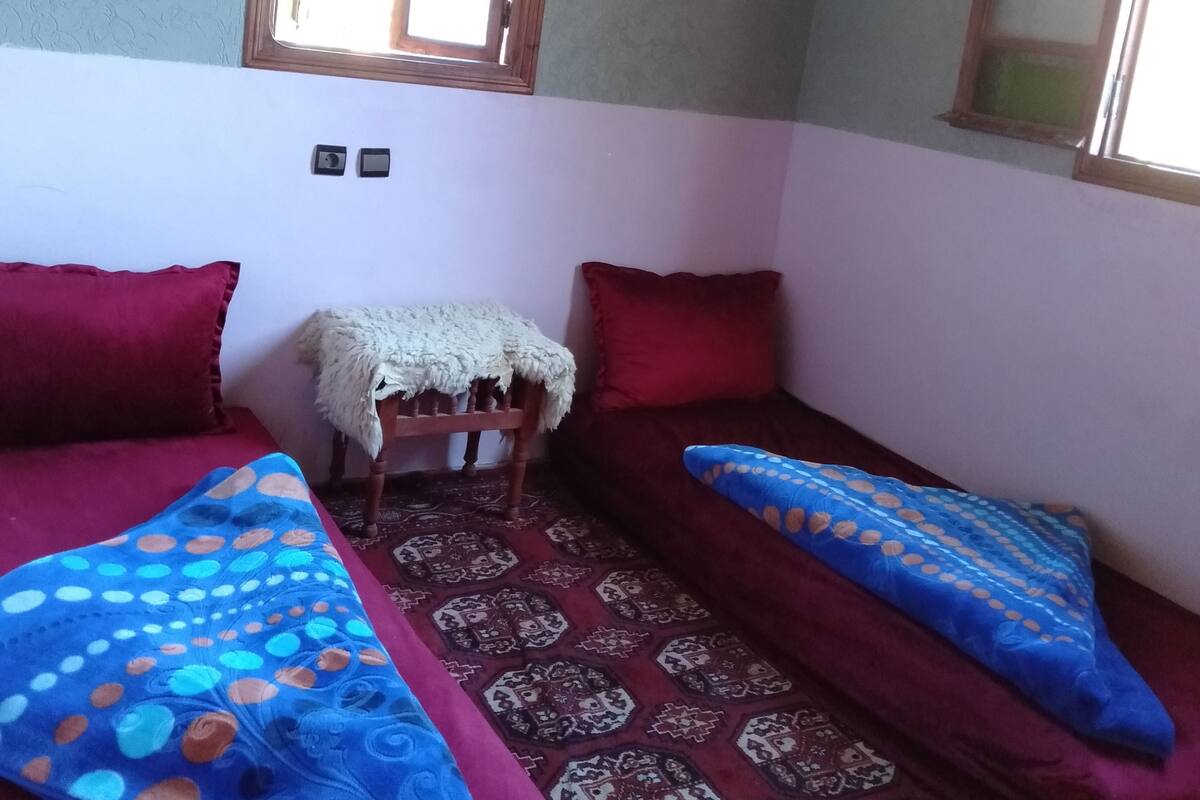 A room with two beds, a small table with a white cloth, patterned rugs, and a window with wooden frames.