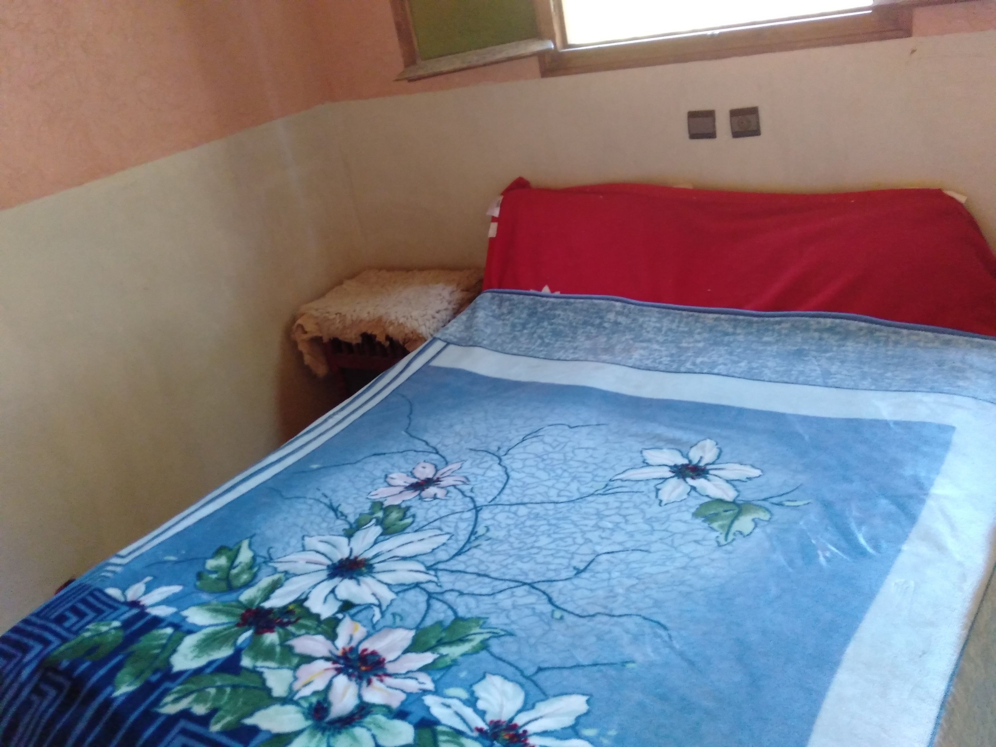 Quarto com cama coberta por um edredom estampado com flores, um travesseiro vermelho e um cobertor bege.