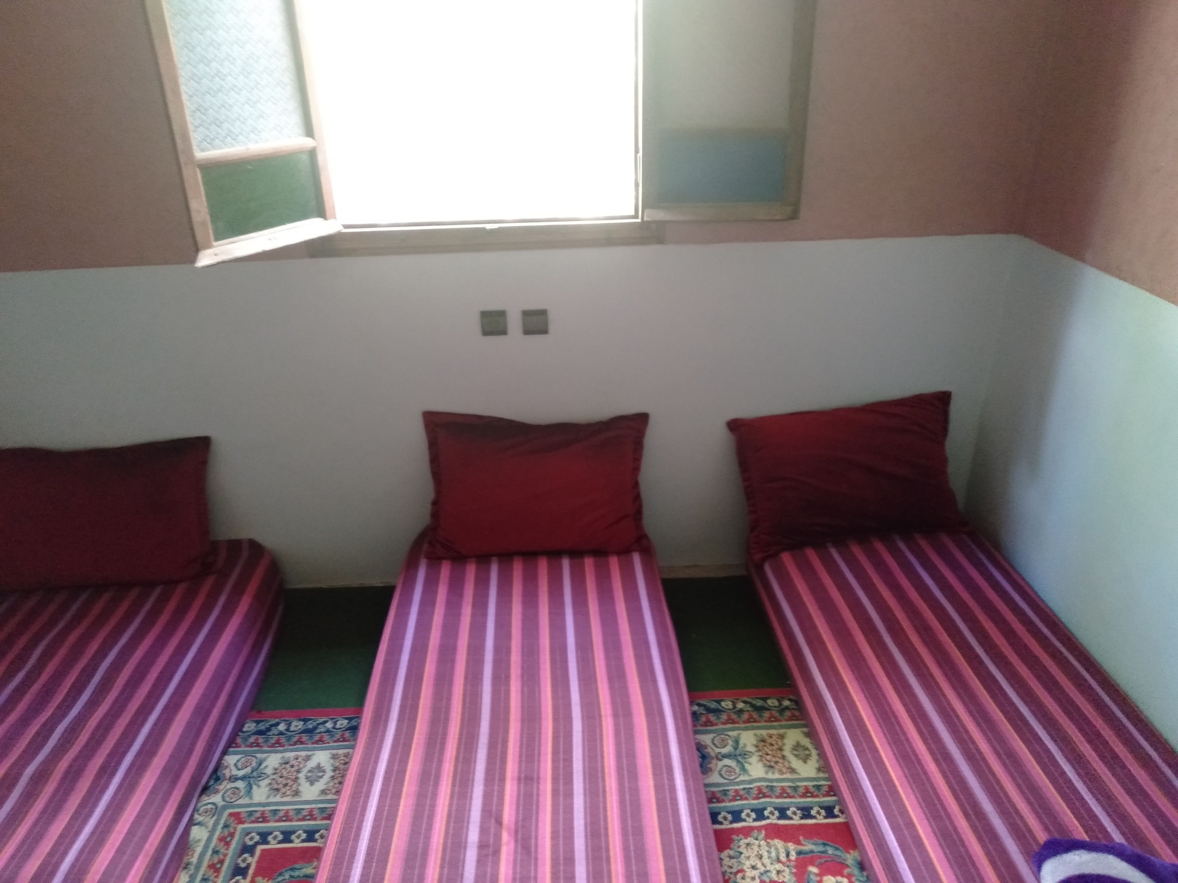 A small room with two single beds, each with a red pillow and a striped bedspread. There is a window above the beds, and the walls are painted in a light color.