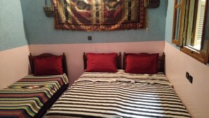 A room with two beds, one with a striped bedspread and the other with a colorful striped bedspread. There is a red pillow on each bed, and a large tapestry hangs on the wall above the beds. - Tanssi House (Asni)
