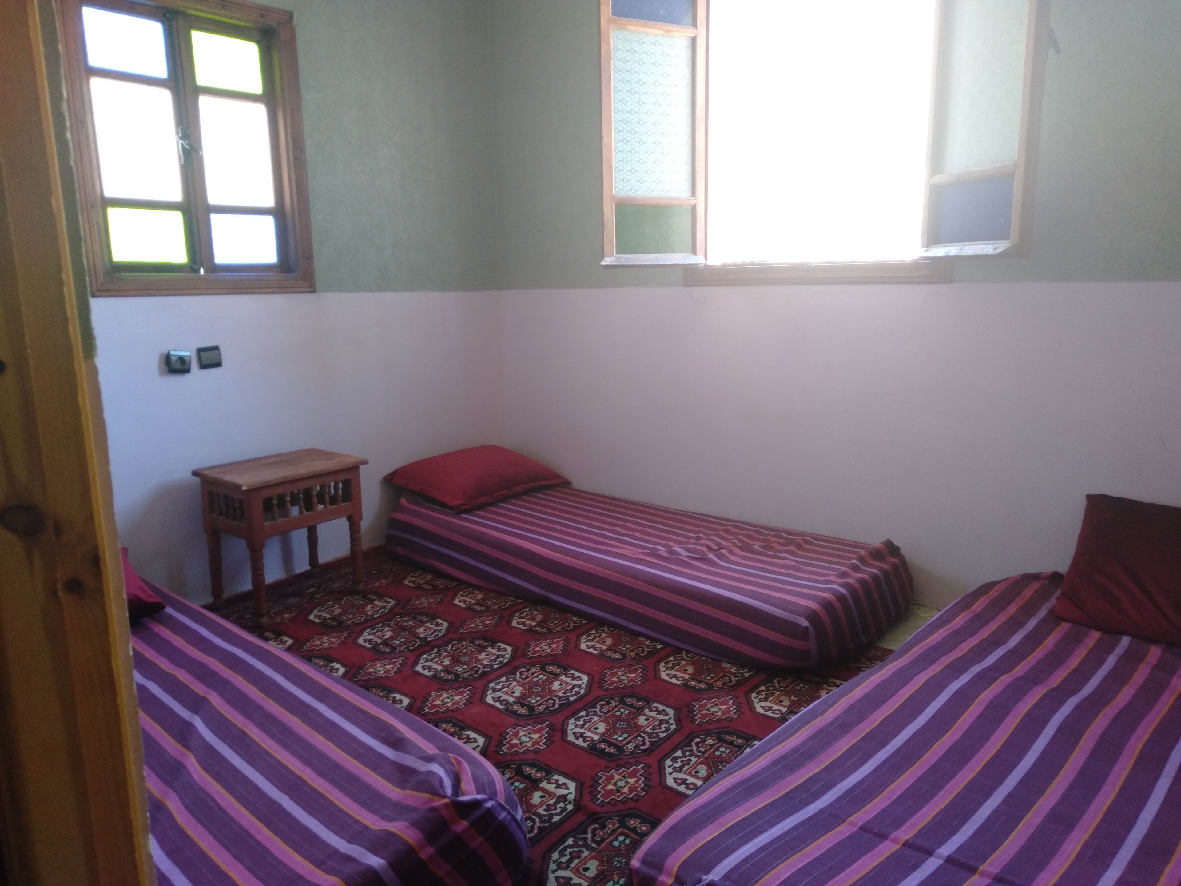 A small, single-room accommodation with two single beds, a small wooden table, and a window providing natural light.