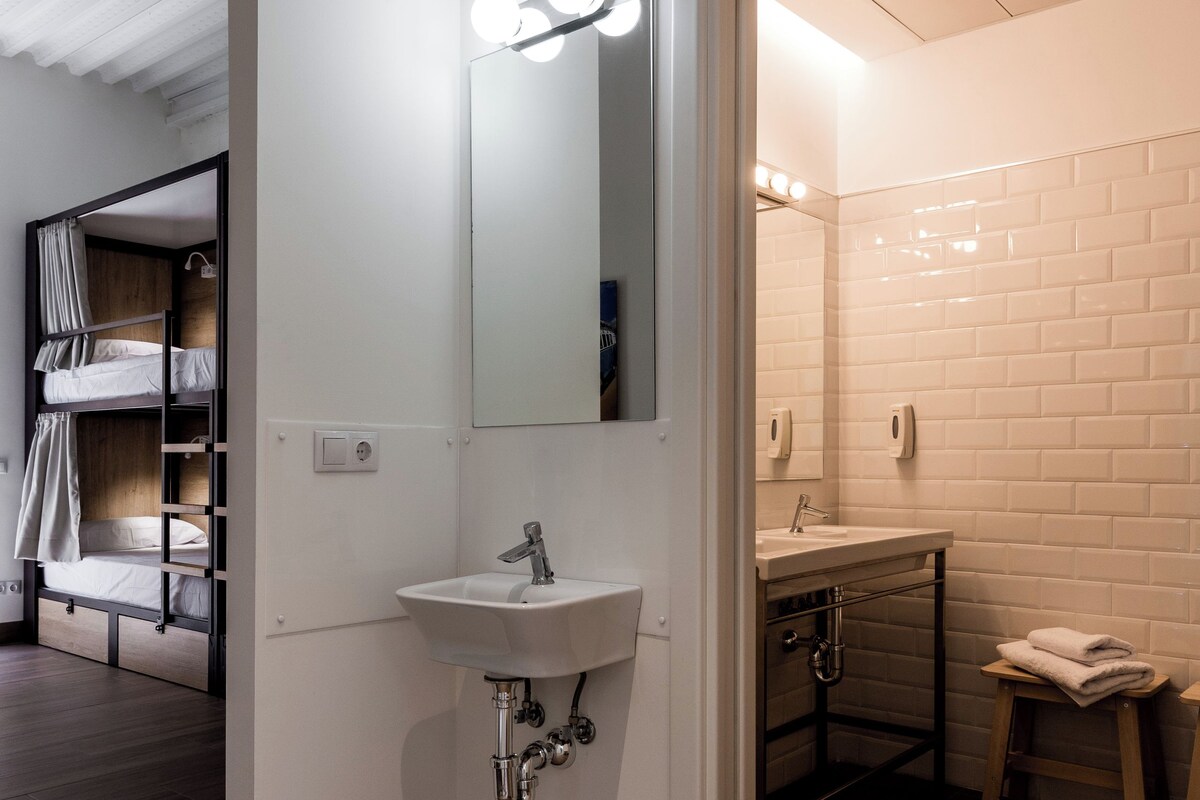 A modern bathroom with a white sink, mirror, and tiled walls. There is a wooden bench and a small stool.