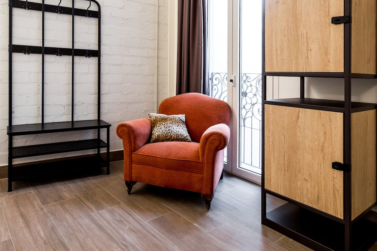 Een kamer met een bakstenen muur, een houten vloer, een rode fauteuil, een zwart metalen plank en een houten kast.