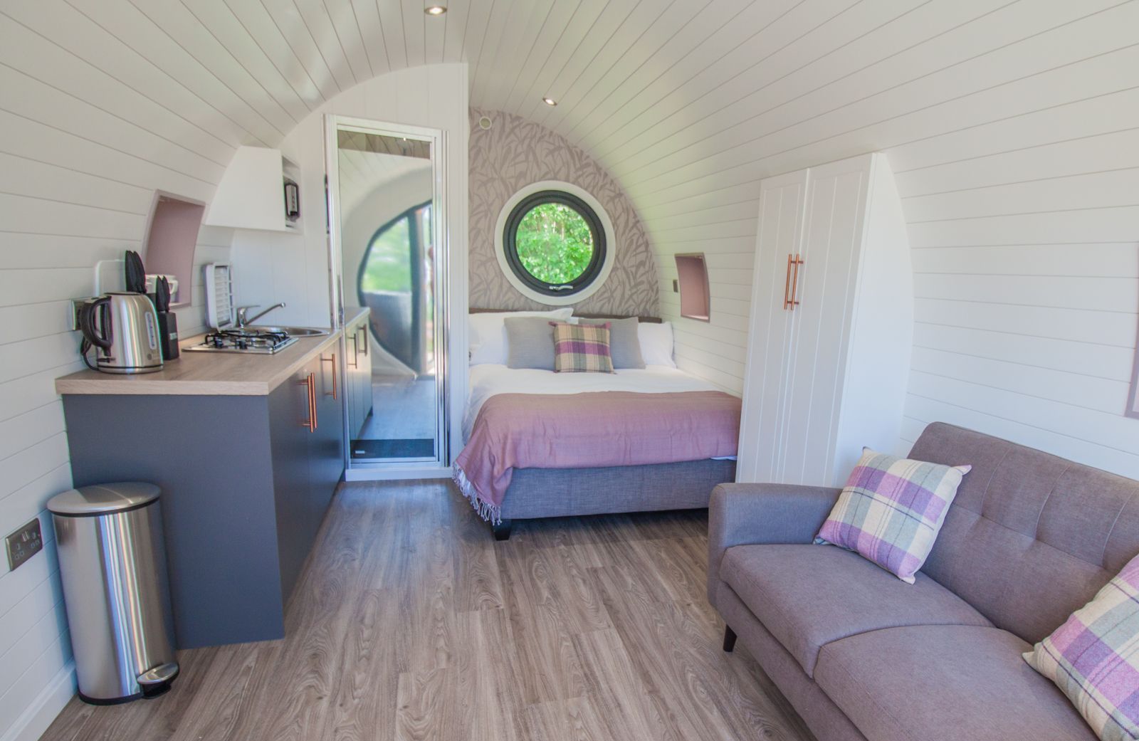 Una camera compatta con un letto, un divano, un angolo cottura e una finestra circolare.