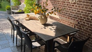 An outdoor patio with a wooden table, black wicker chairs, hanging woven light fixtures, and a brick wall.