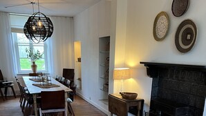 A dining area with a long wooden table, chairs, a hanging light fixture, and a fireplace.