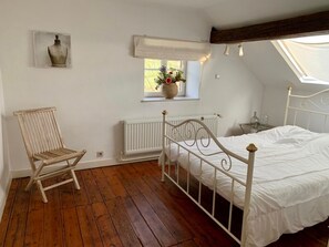 Een slaapkamer met een wit bed, een houten stoel, een radiator, een raam met uitzicht en een plafondlamp.