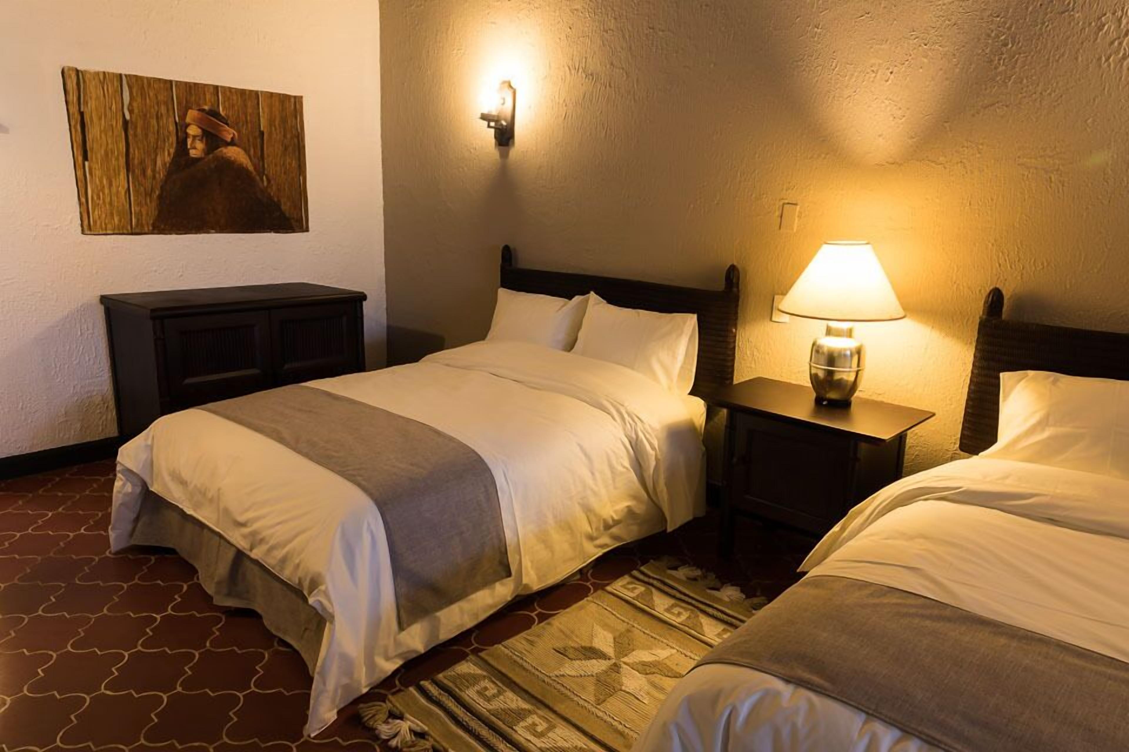 A hotel room with two beds, a nightstand with a lamp, a wall-mounted light, and a painting.
