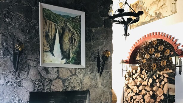A rustic interior with a stone fireplace, a framed painting of a waterfall, and a stack of firewood. - Parador del Alma (Bocoyna)