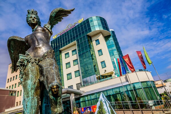 A statue of a winged figure in front of a modern hotel with a glass facade and multiple flags.