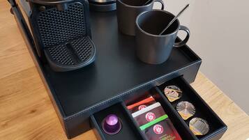 A kitchen counter with a coffee maker, a kettle, and two mugs. Below, a drawer contains a coffee pod, a tea bag, and a small container.