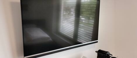 Un televisor de pantalla plana montado en la pared, una cafetera y un control remoto sobre una mesa de madera.