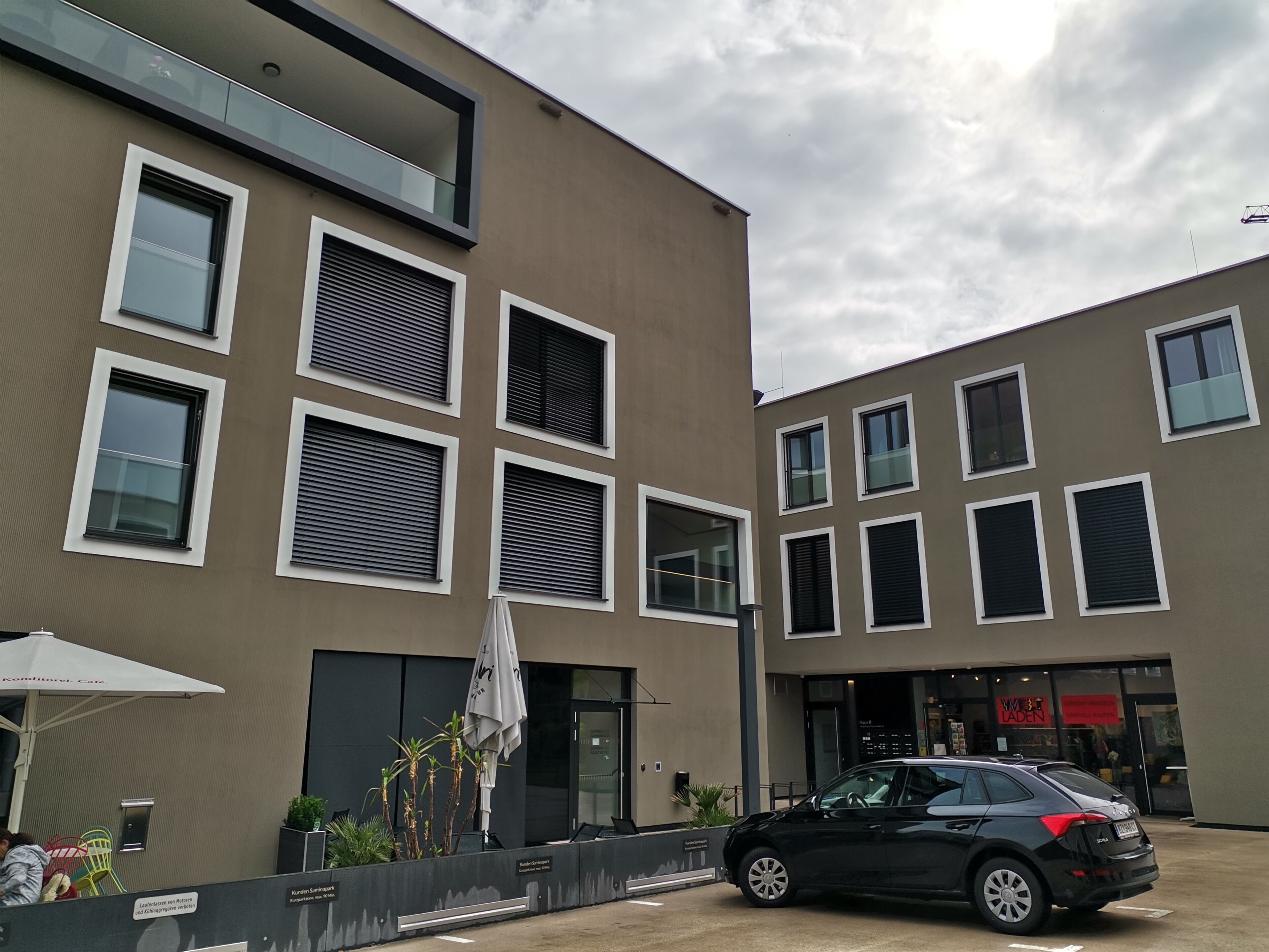 Un edificio moderno con molte finestre, un'auto nera parcheggiata di fronte e posti a sedere all'aperto con ombrelloni.