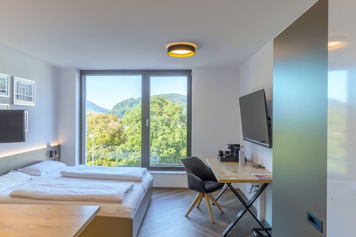 Ein modernes Hotelzimmer mit einem Bett, einem Schreibtisch mit Computer, einem Stuhl und einem großen Fenster mit Blick ins Freie.
