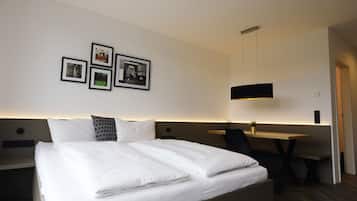 A neatly made bed with white linens, a desk with a chair, and a wall-mounted light fixture.