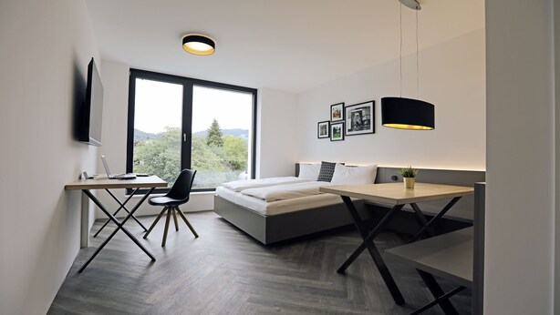 A modern bedroom with a bed, desk, and chair, a TV mounted on the wall, and a window with a view of greenery.