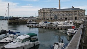 Un puerto deportivo con varias embarcaciones amarradas, un edificio grande con una torre destacada y un paseo marítimo de madera.